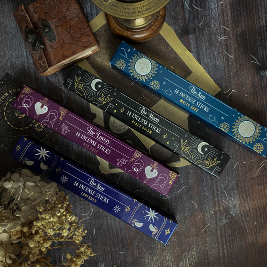 Assortimento di bastoncini d'incenso esoterici con temi astrali e simboli dei tarocchi, come La Luna e La Stella, disposti artisticamente su un tavolo scuro con sfondo di oggetti vintage e botanica essiccata