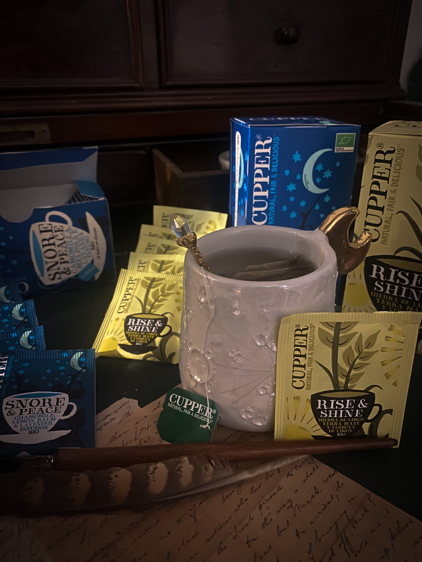 Un momento di relax con una tazza di tisana Cupper, circondata da pacchetti di tè vari assortiti, sullo sfondo di un'ambientazione accogliente con note scritte a mano e una penna stilografica, invitando a una pausa rilassante.