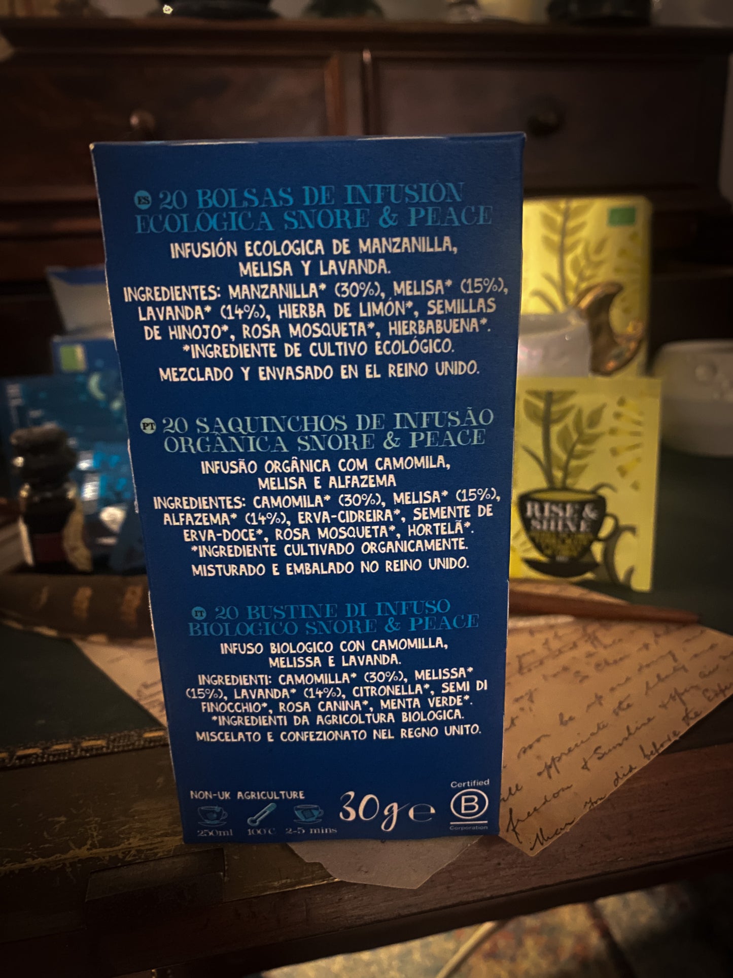 Un momento di relax con una tazza di tisana Cupper, circondata da pacchetti di tè vari assortiti, sullo sfondo di un'ambientazione accogliente con note scritte a mano e una penna stilografica, invitando a una pausa rilassante.