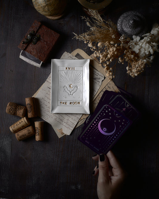Piattino ceramico bianco con incisioni che raffigurano il tarocco 'The Moon', accompagnato da una collezione di carte dei tarocchi color porpora, suggerendo una pratica di riflessione e divinazione. L'ambientazione è arricchita da elementi vintage e una palette di colori caldi e terrosi.