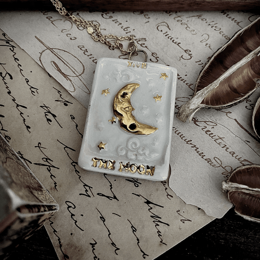 Ciondolo in ceramica raffigurante la luna con dettagli dorati posato su una carta di tarocchi, con penne e un manico di legno sullo sfondo scritto a mano.
