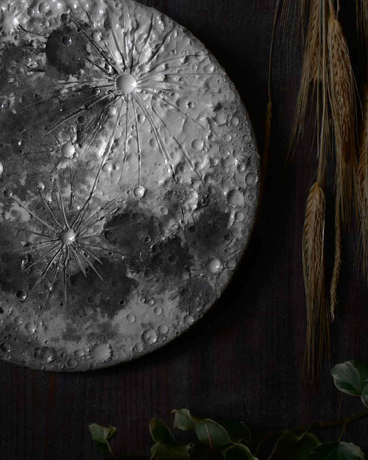 Dettaglio ravvicinato di una decorazione murale in ceramica con texture lunare, appesa su uno sfondo scuro accanto a spighe di grano.