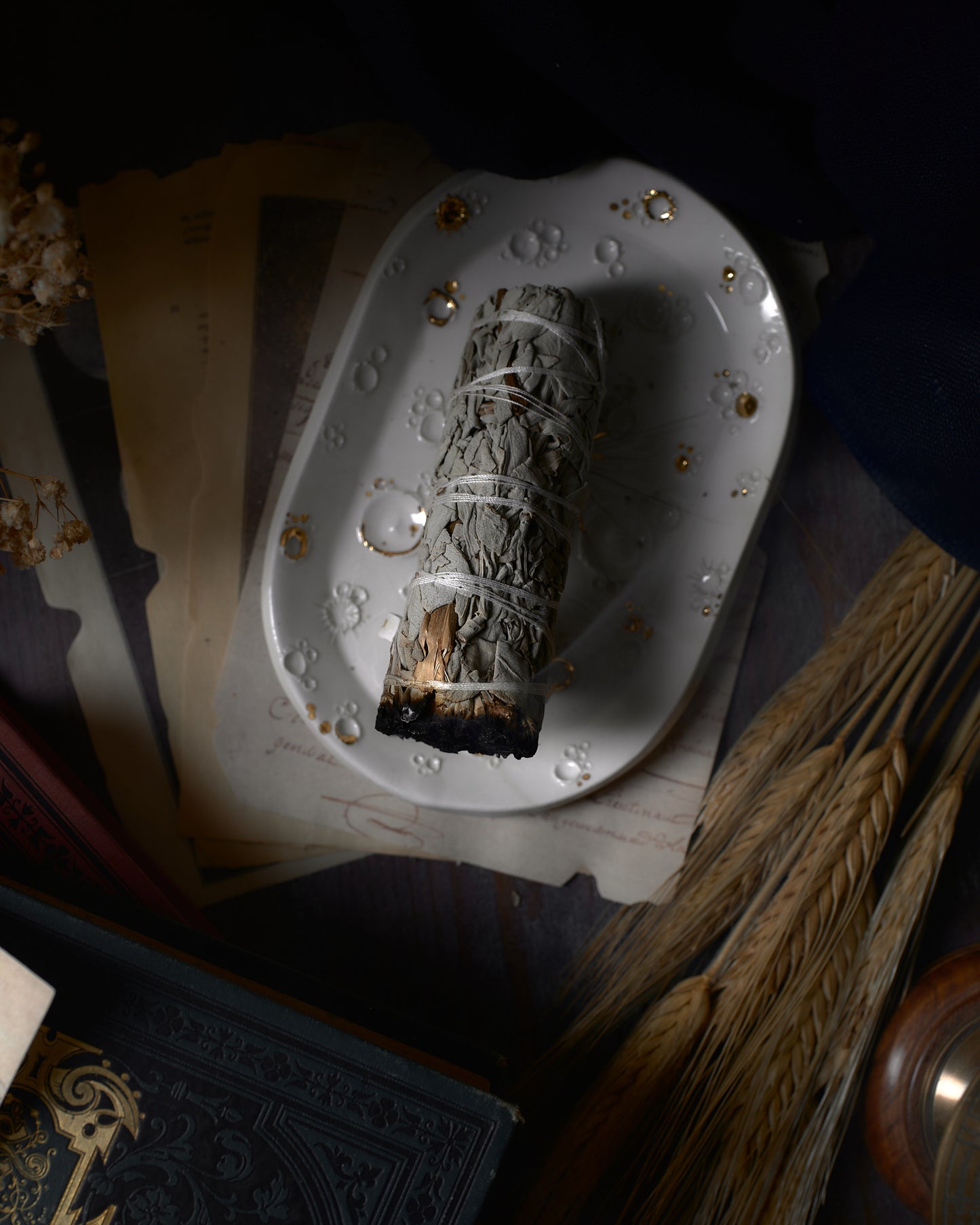 Mazzo di salvia bianca in combustione su portaincenso ceramico bianco con dettagli in oro, con sfondo di oggetti d'epoca e fiori secchi.