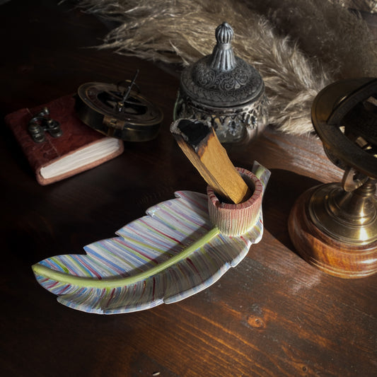 Un portaincenso artistico in ceramica a forma di foglia con una barretta di palo santo, disposto su un tavolo scuro con oggetti vintage come un diario in pelle, un astrolabio e un globo.