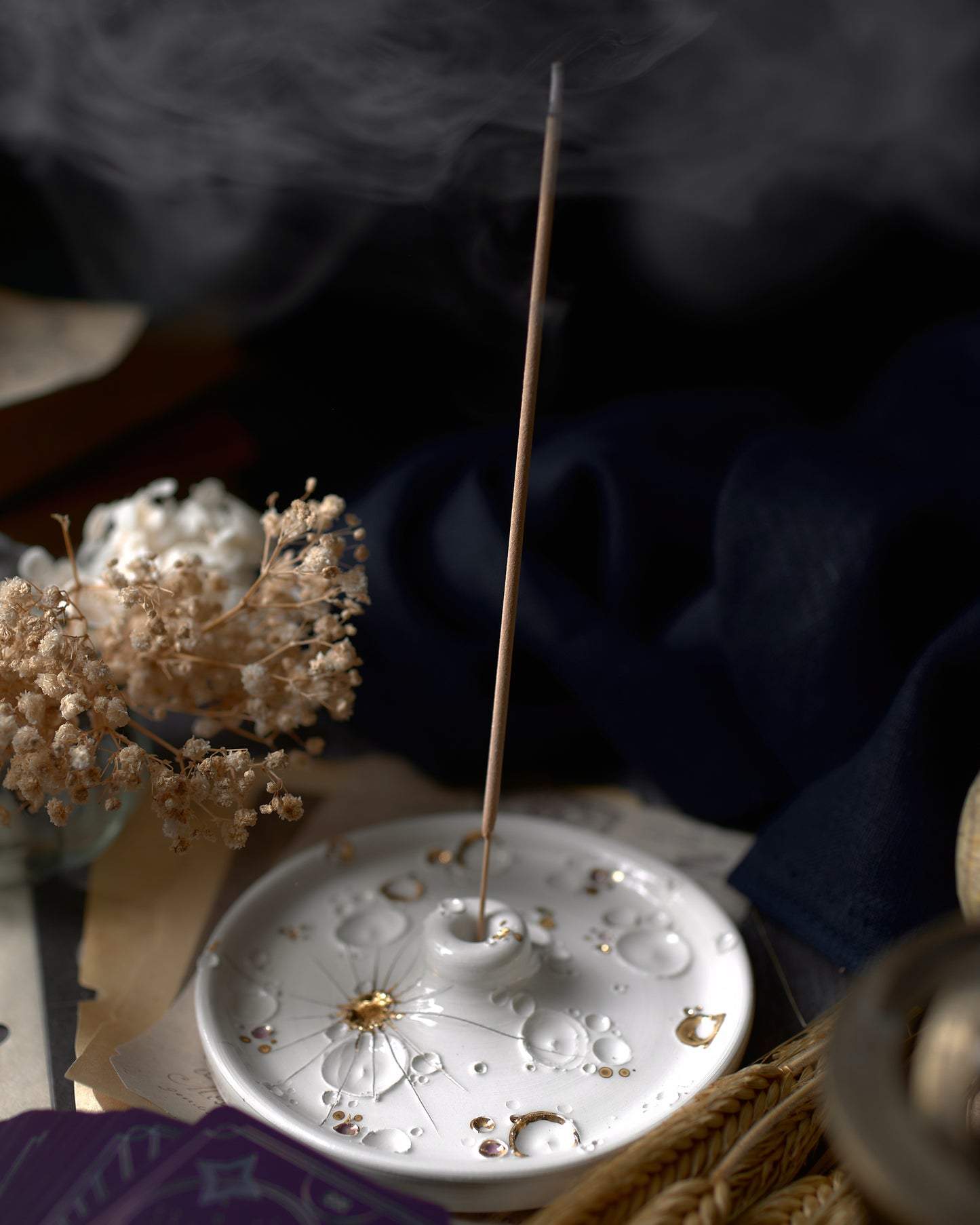 Porta-incenso in ceramica bianca decorato con dettagli dorati e incenso fumante, circondato da un ambiente calmante con fiori secchi e tessuti eleganti.