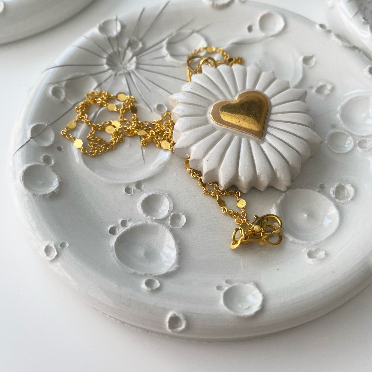Portagioie in ceramica bianca con dettagli dorati, presentando un elegante braccialetto e una collana appoggiati su di esso, in una composizione pura e sofisticata.