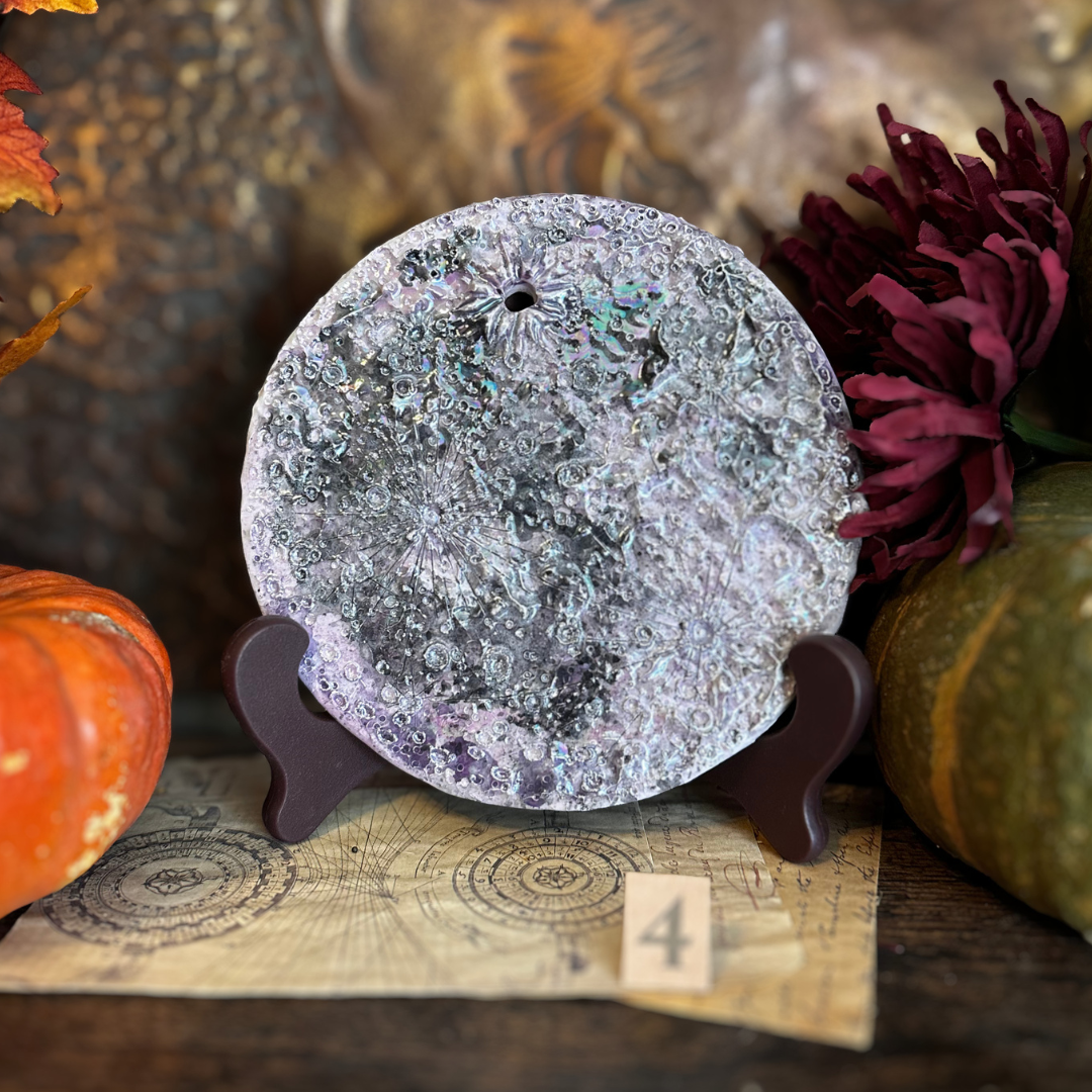 Luna della Soglia IV, luna media in ceramica viola su un leggio con il numero 4, fra zucche e fiori scuri