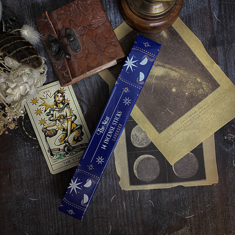 Bastoncini d'incenso esoterici con temi astrali e simboli dei tarocchi, come La Luna e La Stella, disposti artisticamente su un tavolo scuro con sfondo di oggetti vintage e botanica essiccata
