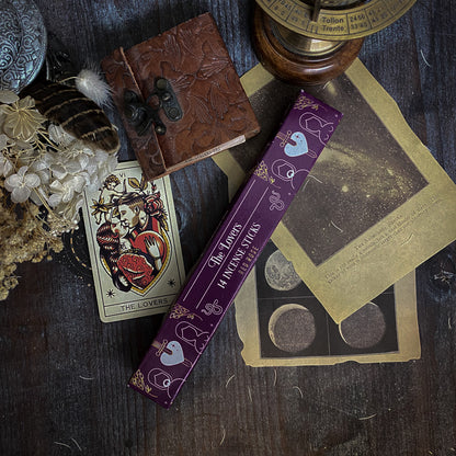 Bastoncini d'incenso esoterici con temi astrali e simboli dei tarocchi, come La Luna e La Stella, disposti artisticamente su un tavolo scuro con sfondo di oggetti vintage e botanica essiccata