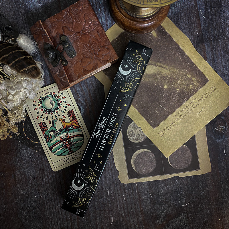 Bastoncini d'incenso esoterici con temi astrali e simboli dei tarocchi, come La Luna e La Stella, disposti artisticamente su un tavolo scuro con sfondo di oggetti vintage e botanica essiccata