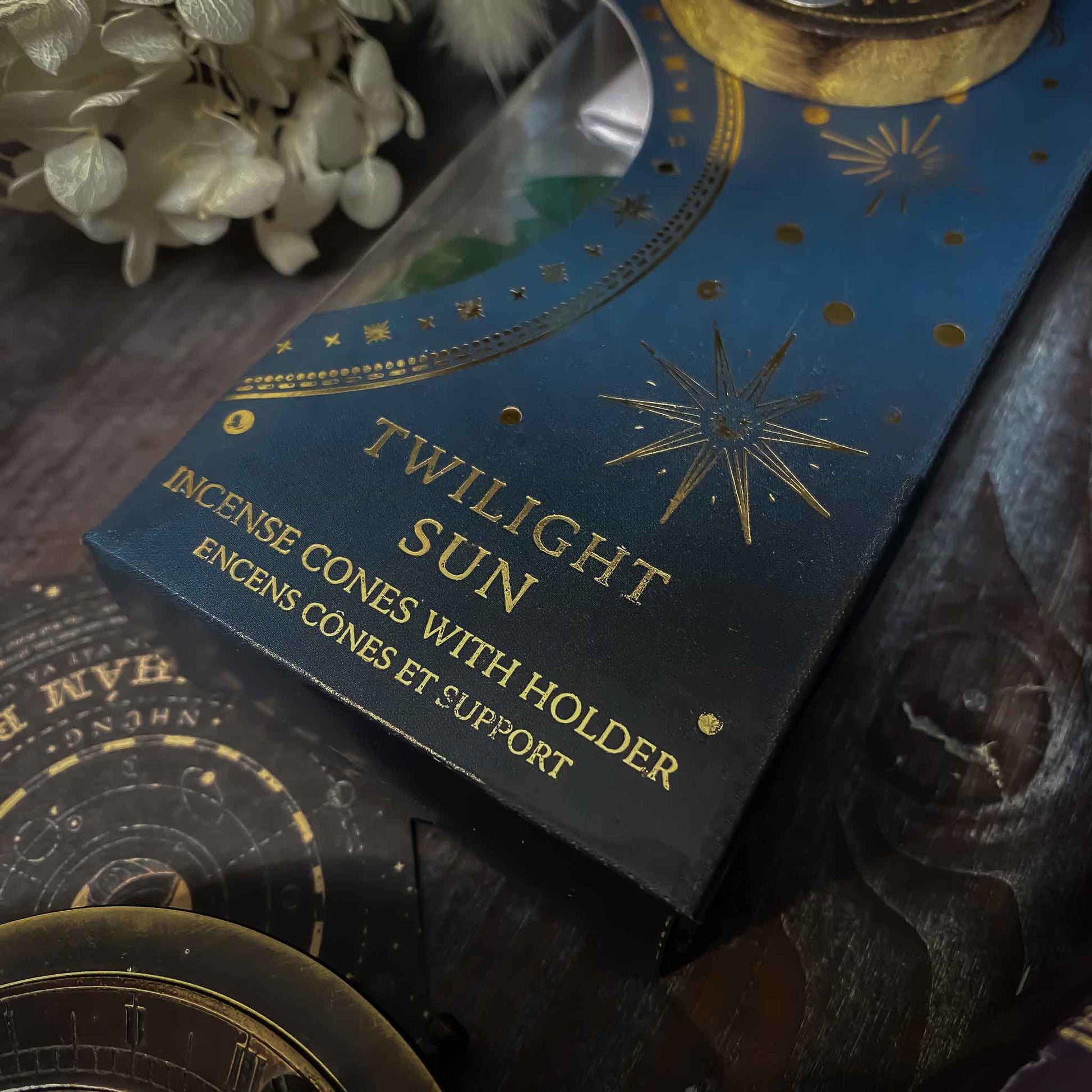 incenso twilight sun, Assortimento di coni di incenso con supporto dalle fragranze celesti, ciascuno in una confezione tematica con motivi del Sole e della Luna, disposti artisticamente su un tavolo in legno, accanto a oggetti vintage e fiori secchi, evocando un'atmosfera di tranquillità e introspezione.