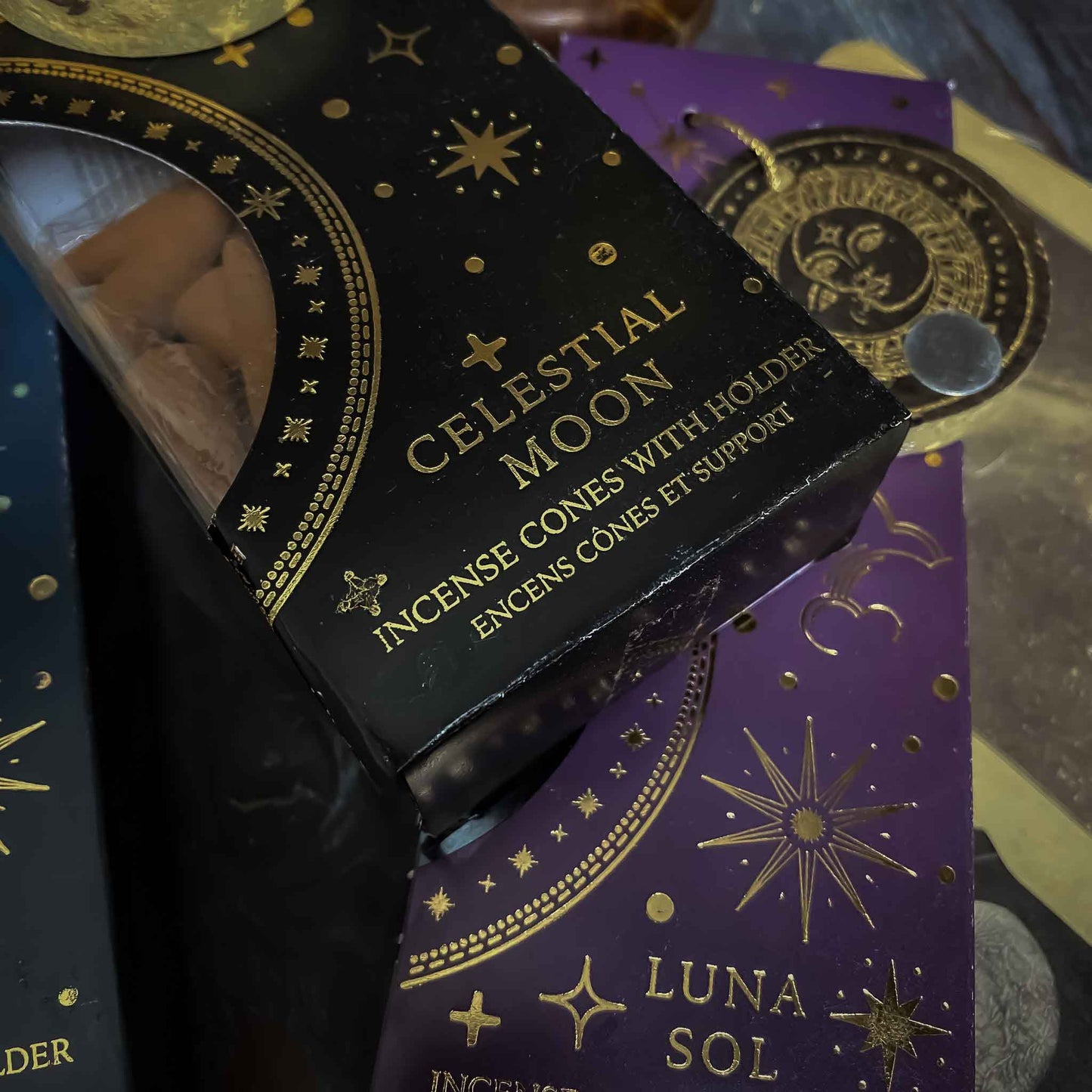incenso celestial moon, Assortimento di coni di incenso con supporto dalle fragranze celesti, ciascuno in una confezione tematica con motivi del Sole e della Luna, disposti artisticamente su un tavolo in legno, accanto a oggetti vintage e fiori secchi, evocando un'atmosfera di tranquillità e introspezione.