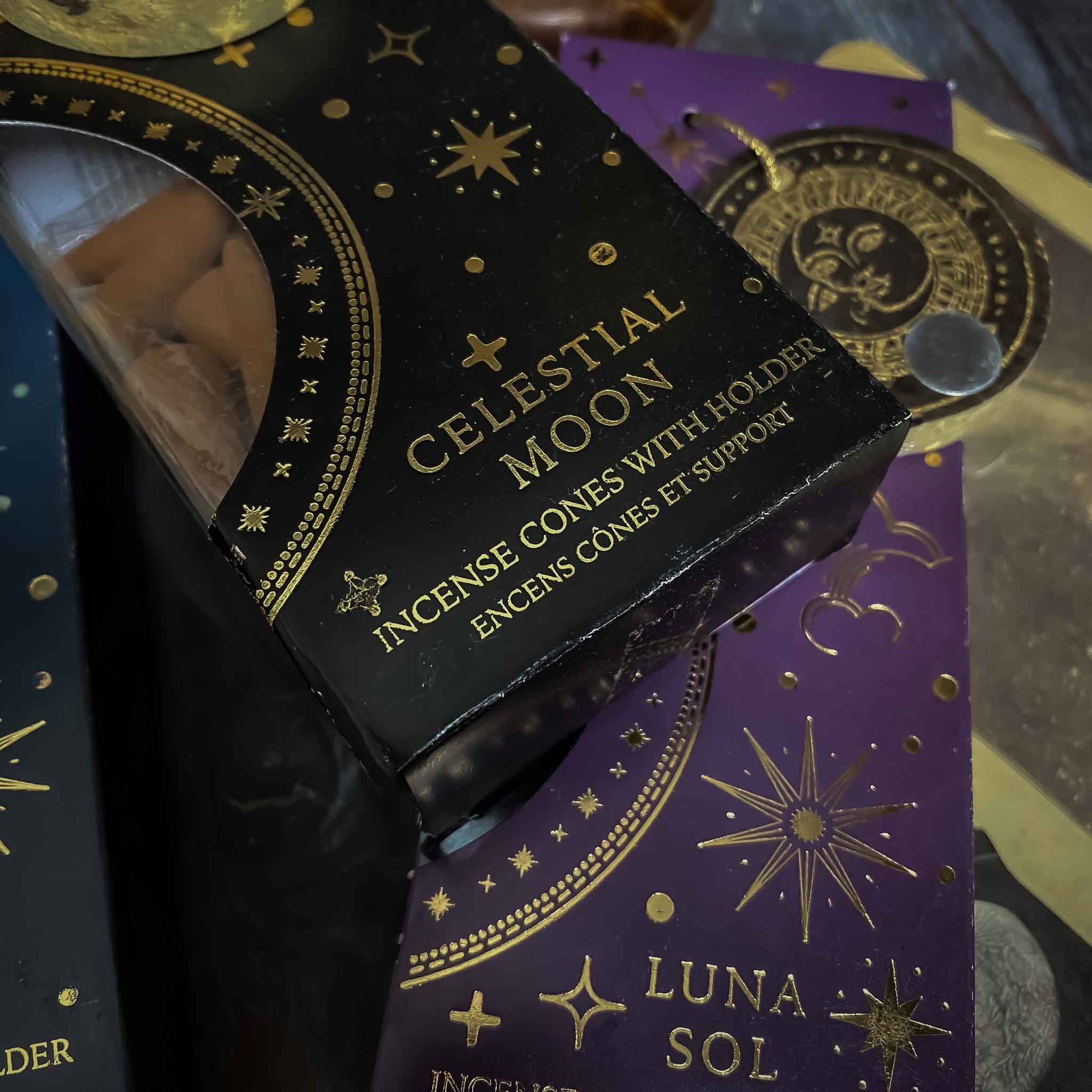incenso celestial moon, Assortimento di coni di incenso con supporto dalle fragranze celesti, ciascuno in una confezione tematica con motivi del Sole e della Luna, disposti artisticamente su un tavolo in legno, accanto a oggetti vintage e fiori secchi, evocando un'atmosfera di tranquillità e introspezione.