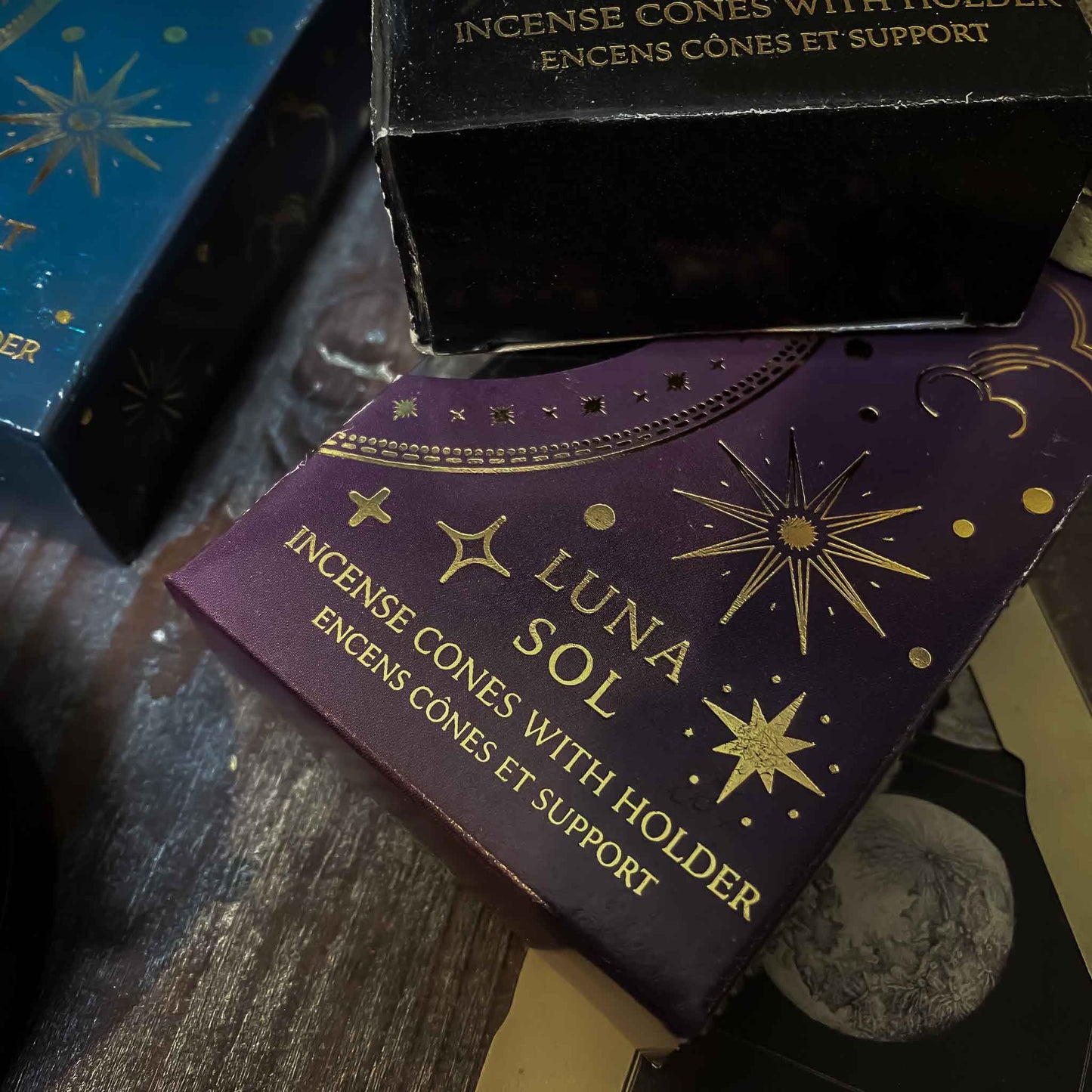 incenso luna sol. Assortimento di coni di incenso con supporto dalle fragranze celesti, ciascuno in una confezione tematica con motivi del Sole e della Luna, disposti artisticamente su un tavolo in legno, accanto a oggetti vintage e fiori secchi, evocando un'atmosfera di tranquillità e introspezione.