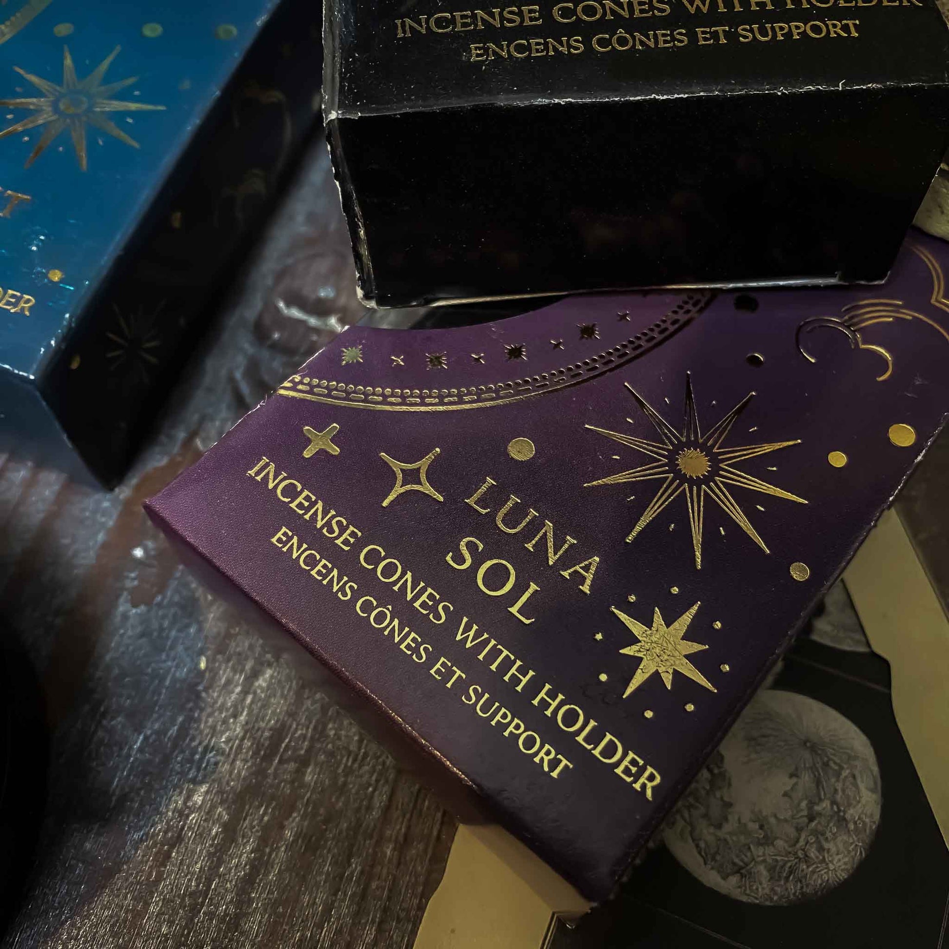 incenso luna sol. Assortimento di coni di incenso con supporto dalle fragranze celesti, ciascuno in una confezione tematica con motivi del Sole e della Luna, disposti artisticamente su un tavolo in legno, accanto a oggetti vintage e fiori secchi, evocando un'atmosfera di tranquillità e introspezione.