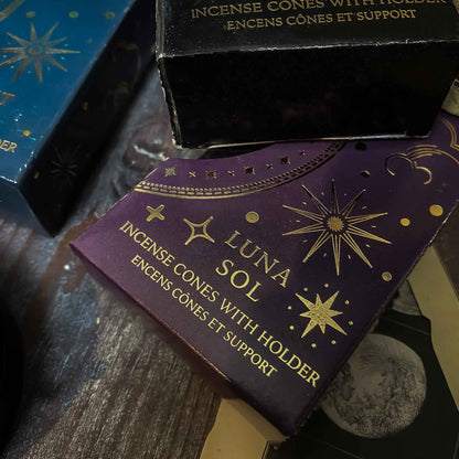 incenso luna sol. Assortimento di coni di incenso con supporto dalle fragranze celesti, ciascuno in una confezione tematica con motivi del Sole e della Luna, disposti artisticamente su un tavolo in legno, accanto a oggetti vintage e fiori secchi, evocando un'atmosfera di tranquillità e introspezione.