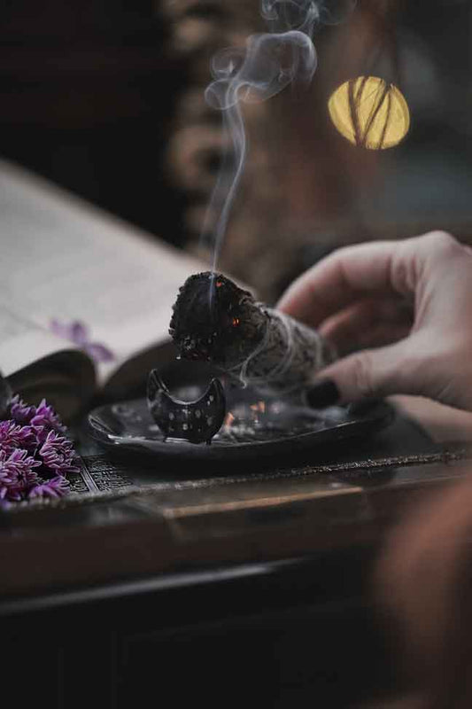 MANO CHE APOGGIA LA SALVIA CHE STA BRUCIANDO SU UN PIATTINO NERO LUNARE