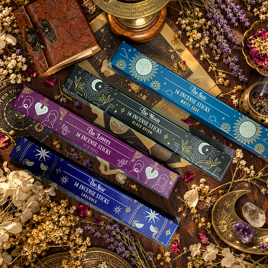 Assortimento di bastoncini d'incenso esoterici con temi astrali e simboli dei tarocchi, come La Luna e La Stella, disposti artisticamente su un tavolo scuro con sfondo di oggetti vintage e botanica essiccata
