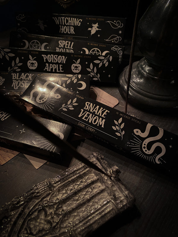 l'immagine presenta una scatola di incensi denominata "Snake Venom", posata tra altre confezioni dal design tematico. In primo piano si trova un incensiere con un bastoncino di incenso acceso, e una scultura che aggiunge un tocco di antico mistero. L'illuminazione è soffusa, contribuendo all'atmosfera occulta e intima. Il design del packaging è caratterizzato da simboli bianchi contrastanti sullo sfondo nero, creando un look distintivo ed elegante.