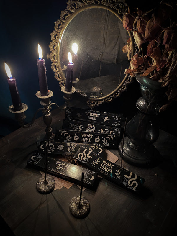 Nell'immagine è visibile un'ampia varietà di confezioni di incensi, con in evidenza i nomi "Black Rose" e "Midnight Moon" tra gli altri. Le scatole sono disposte in maniera strategica su un tavolo di legno scuro, con incensi accesi inseriti in portaincensi artistici, mentre sullo sfondo si scorgono dei candelabri che conferiscono un'aura gotica e un senso di tradizione.