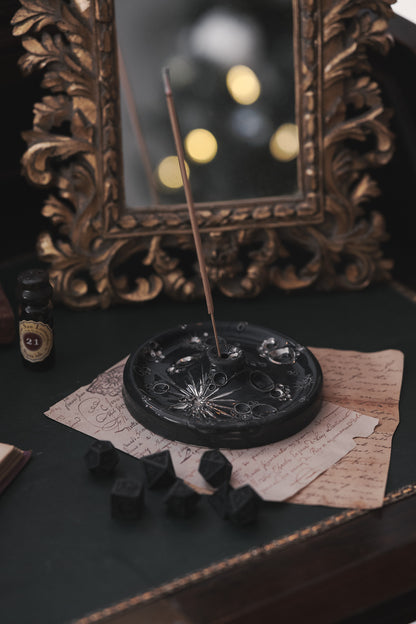 Testo alternativo: Porta-incenso lunare in ceramica posato su un tavolo, con dadi neri e carte manoscritte in primo piano e uno specchio con cornice dorata sfocato sullo sfondo.
