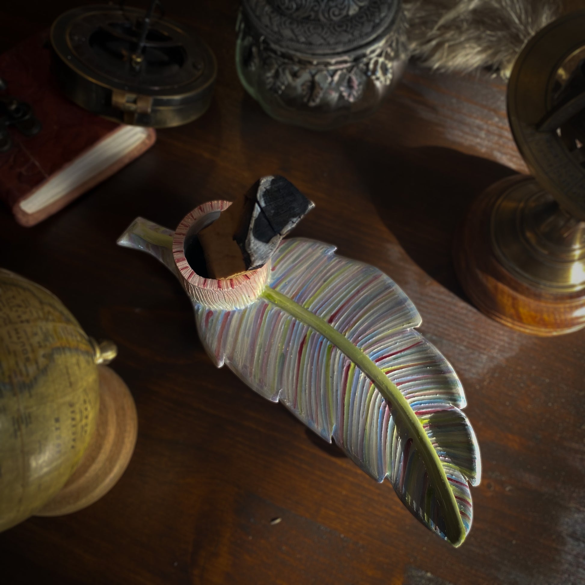 Un portaincenso artistico in ceramica a forma di foglia con una barretta di palo santo, disposto su un tavolo scuro con oggetti vintage come un diario in pelle, un astrolabio e un globo.