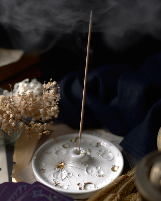 Porta-incenso in ceramica bianca decorato con dettagli dorati e incenso fumante, circondato da un ambiente calmante con fiori secchi e tessuti eleganti.