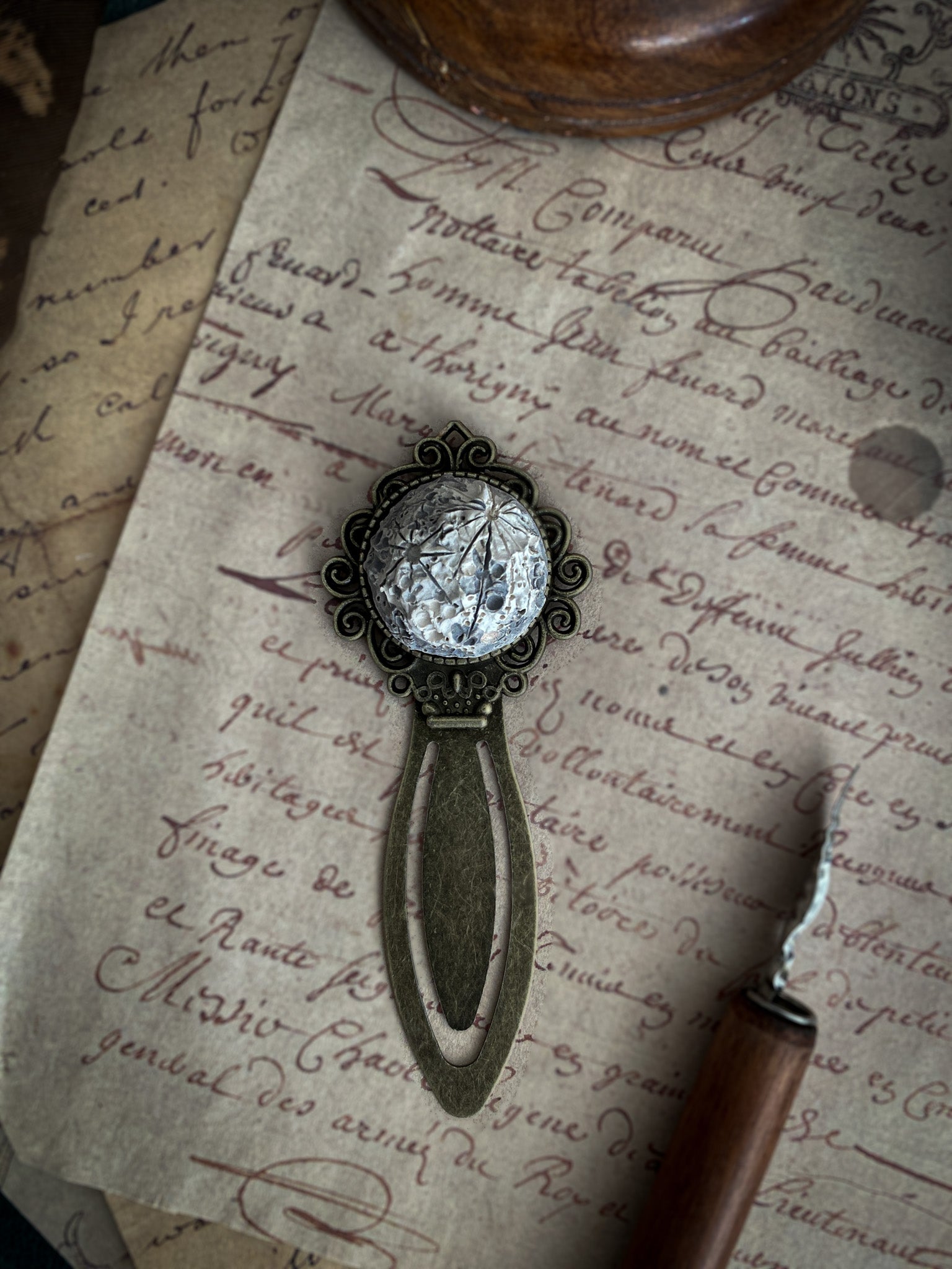 Segnalibro in ceramica con texture lunare posato su carta antica, accanto a un pennino d'inchiostro, invitando all'immersione nella lettura.