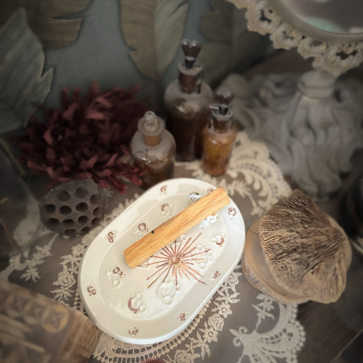 Vista del piattino White and Rose Gold con palo santo, circondato da pizzo, fiori secchi e boccette.