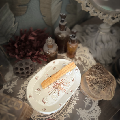 Vista del piattino White and Rose Gold con palo santo, circondato da pizzo, fiori secchi e boccette.